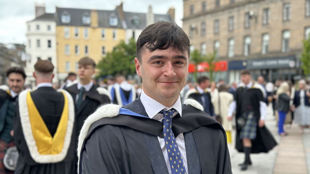 Young male with dark hair wearing graduation gown and hood, with suit and tie underneath. Others in hoods and gowns in background