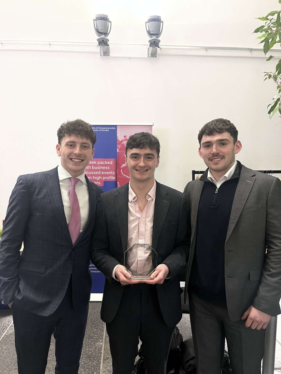 Three young men wearing suits and ties. Male in centre holds a glass trophy.