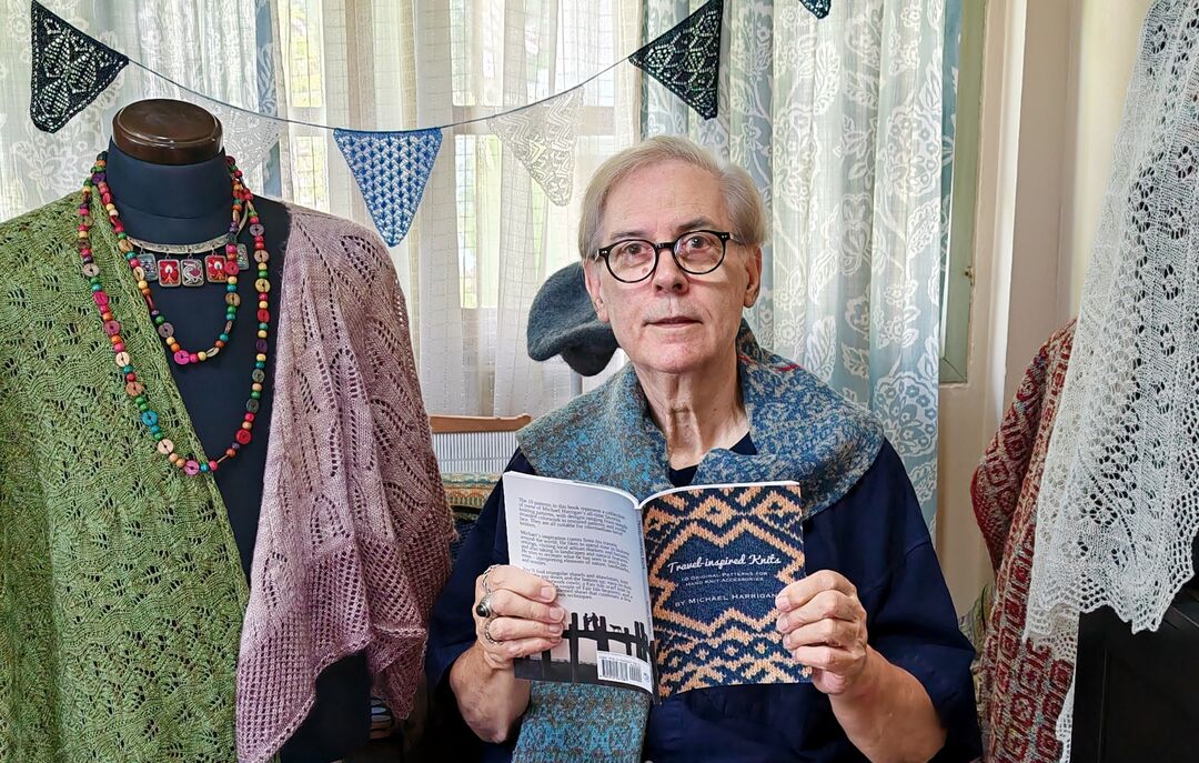 Man wearing a knitted scarf and holding a book, looking at the camera. Surrounded by knitted materials, including a cardigan on a clothing dummy