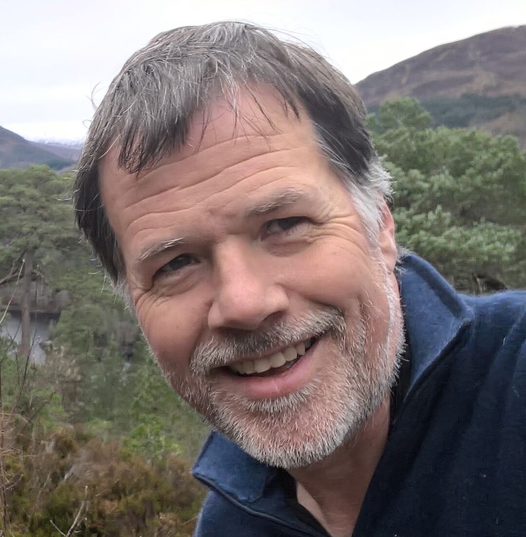 Close up image of man looking straight at camera and smiling. Older man with greying hair and outside in the countryside