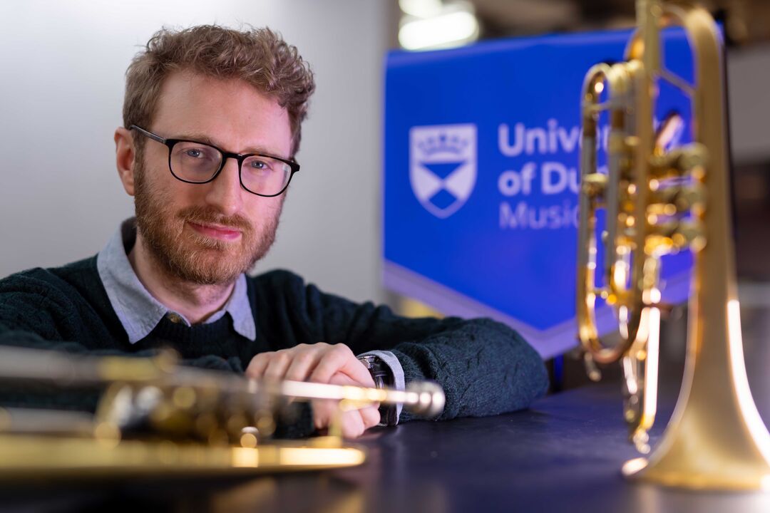 One trumpet stands on it's end to the right, and another lies across a table in the foreground. A blue banner is in the background that says University of Dundee music. A man with glasses sits in the middle.