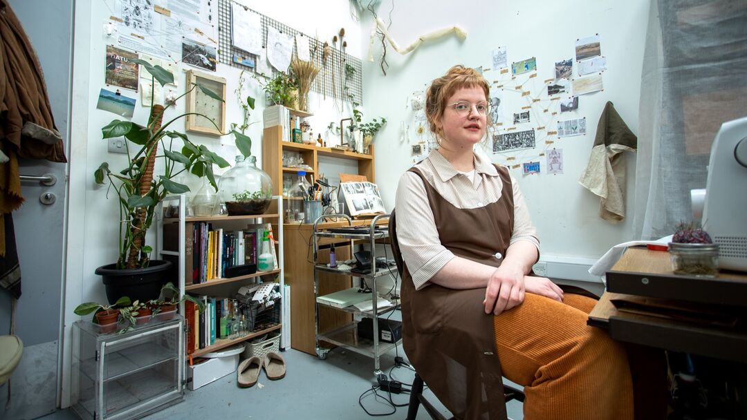 woman sitting off centre on a stool looking towards camera in room with has lots and lots of framed pictures and images on the walls behind