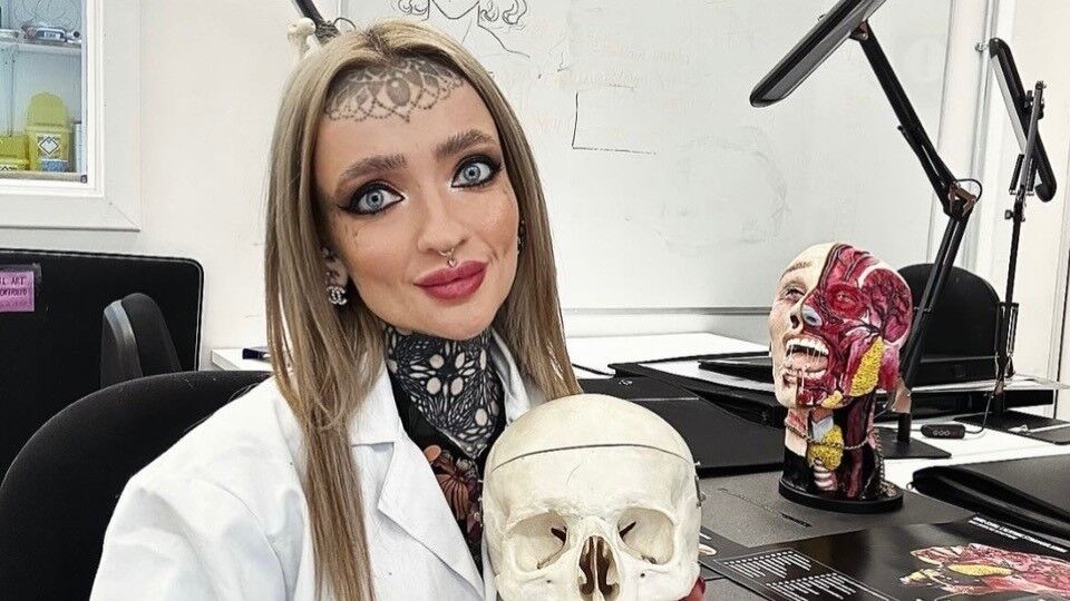 Young woman with long, straight blond hair and a tattoo on her forehead, wearing a white lab coat and holding a skull