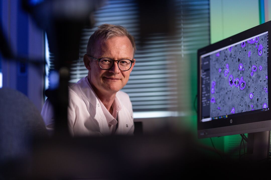 Man wearing glasses and whit lab coat looking towards the camera. Next to him is a screen with science cells on it. the image features blue and green lighting