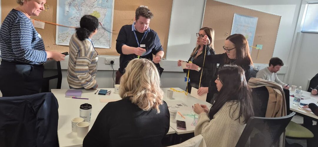 group of people around a table, some sitting and some standing. A few are holding long pieces of string. one is looking at a map on a wall behind her and holding up some string to the map.