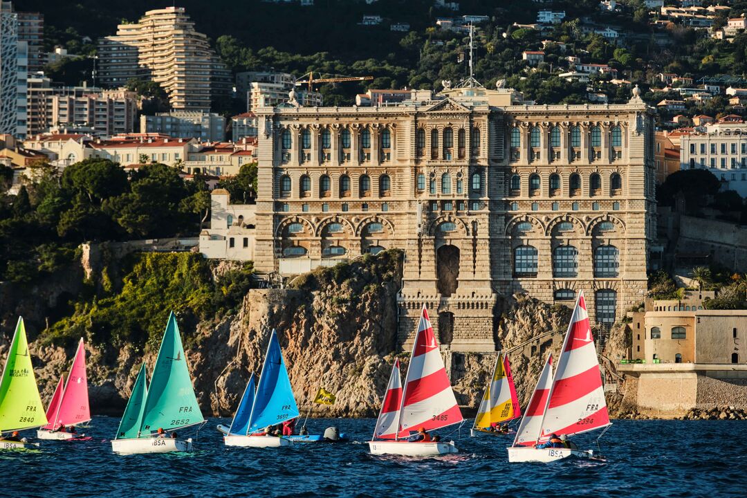 Several colourful sail boats taking part of the Navicap Challenge in Monaco with a stately building on shore in the background