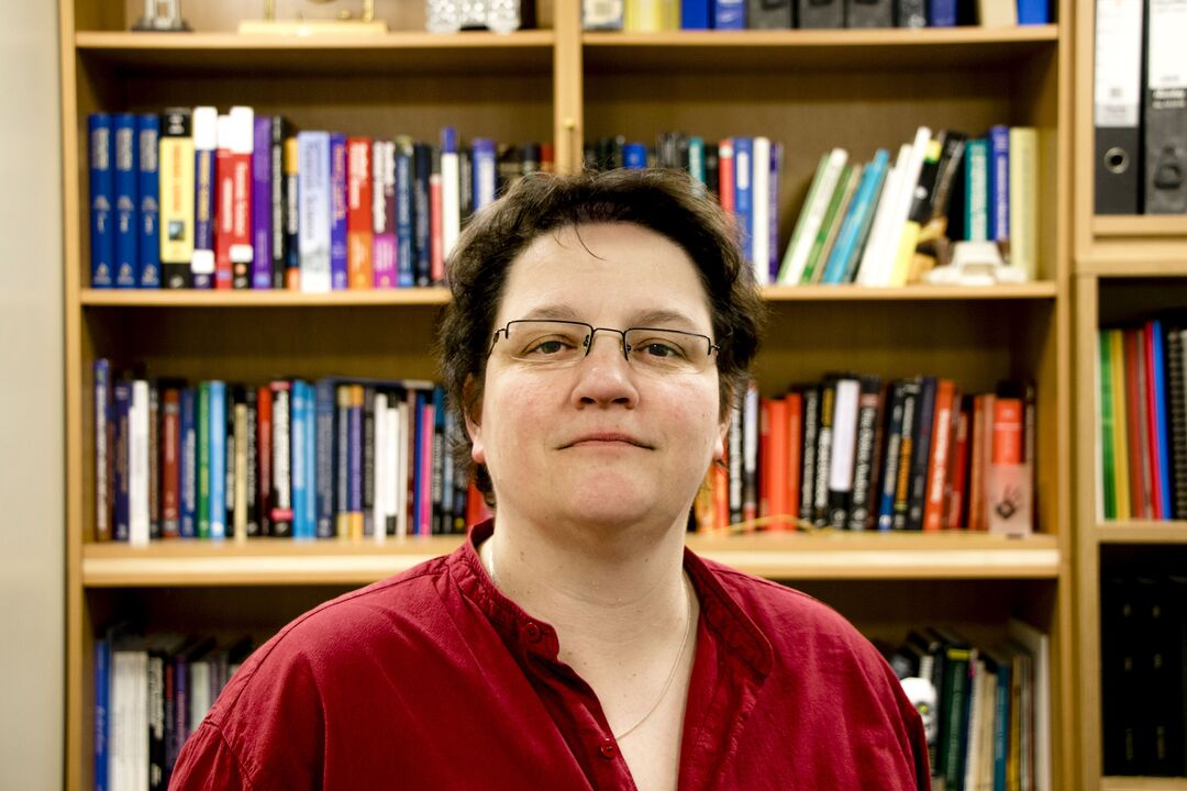 Woman with very short dark hair and glasses standing in front of a bookcase filled with books