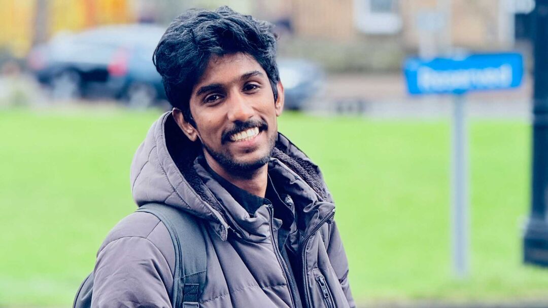 Gokul Raghunathan wearing a large grey coat and backpack, standing outside. There is grass and a parked car in the background.