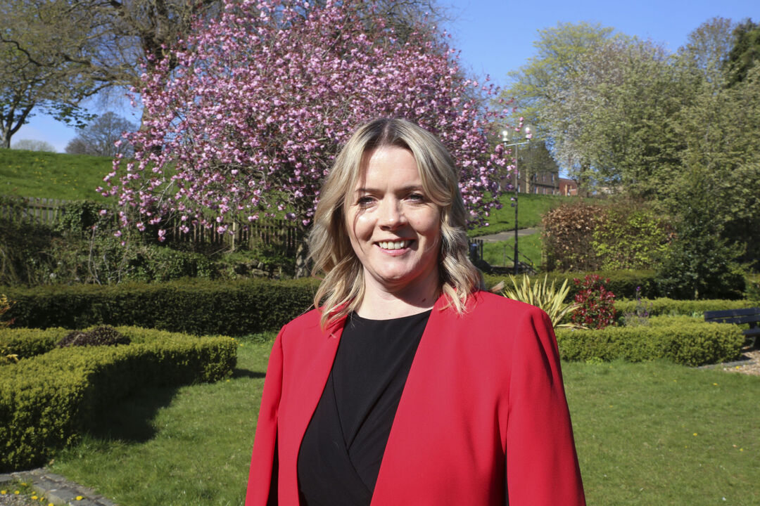 Kirsty Thomson wearing a bright red blazer, standing in a well tended garden with a pink blossoming tree behind her.