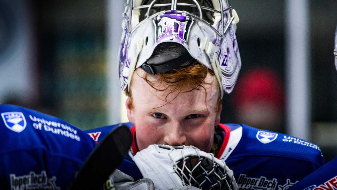 A person wearing a hockey jersey with a helmet pushed up on his head, holds his glove to his mouth.