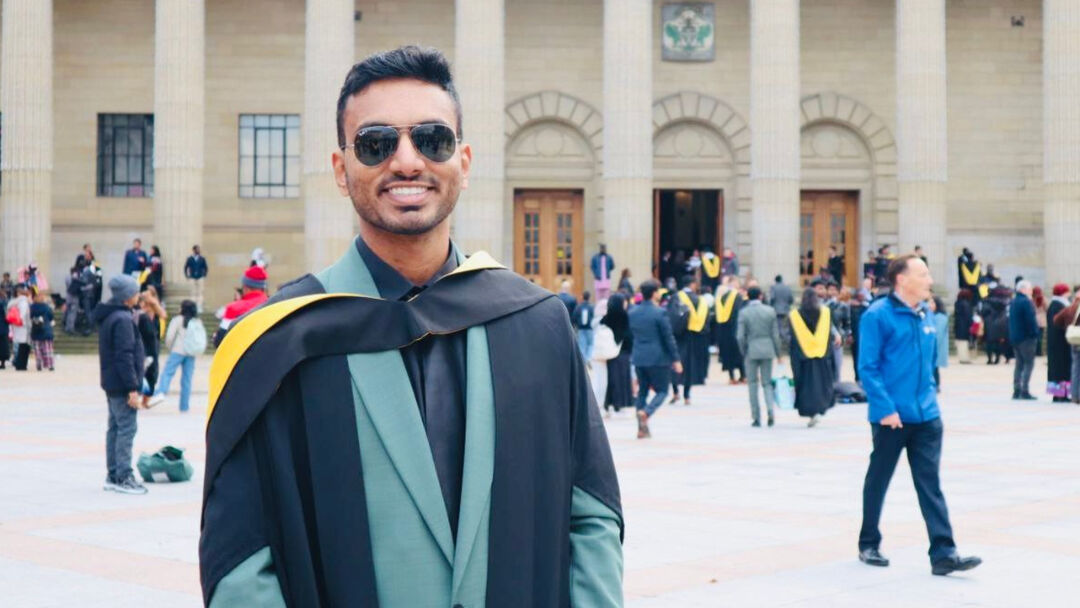 A student poses and smiles at the camera on graduation day, wearing a green suite with black shirt and graduation gown