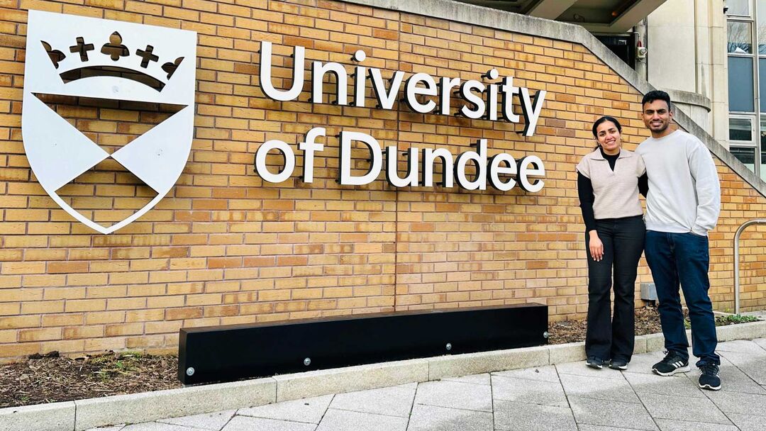 A young couple posing together infront of a University of Dundee sign, male wearing a grey jumper, blue jeans and female wearing a cream jumper and black jeans.