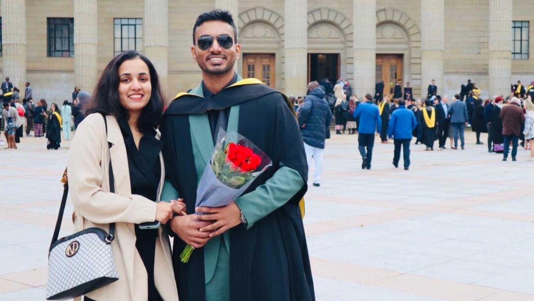A young couple posing together on graduation day, male wearing a green suit and graduation gown, with sunglasses, and female wearing a cream jumper, black top and a handbag over her shoulders, with graduates in the background.