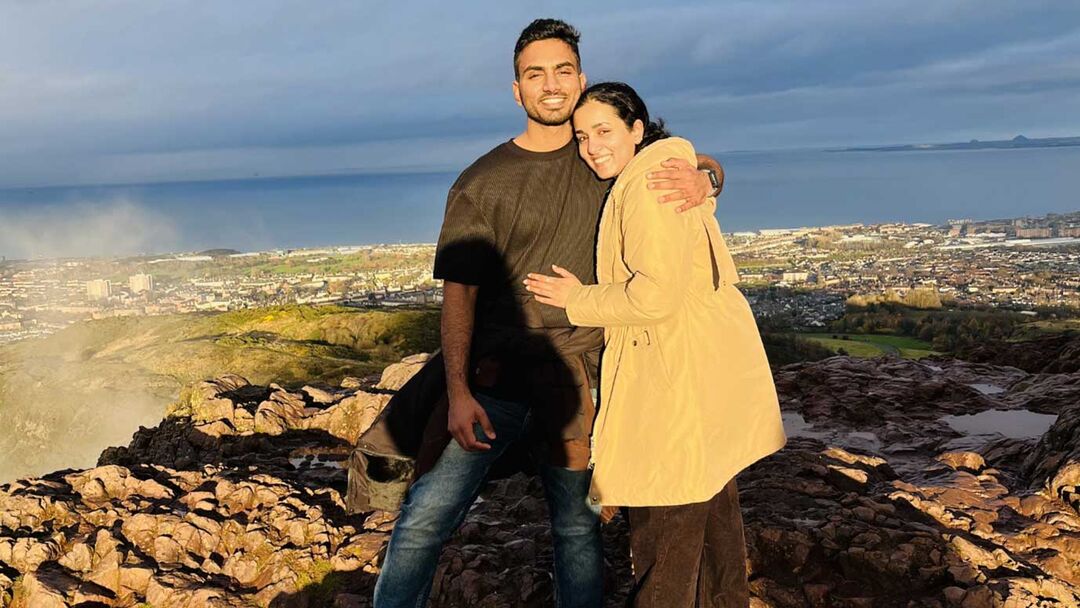 A young couple posing together at the top of Arthurs seat, male wearing a grey tshirt and blue jeans and female wearing a cream jumper jacket with the city and sky in the background.