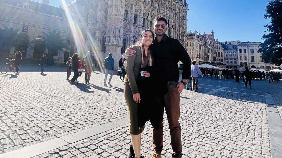 A young couple posing together male wearing a black top and tan trousers and female wearing a green jumper/dress outside with the sun shinning.