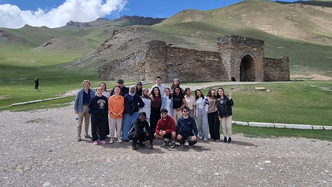 A large group of students, standing in a line smiling at the camera, standing outside with a mountains behind and an old building