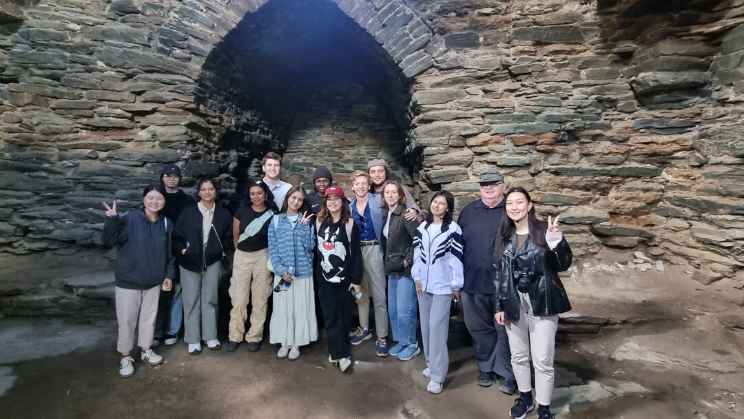 A large group of students, standing in a line smiling at the camera, standing inside a ruin with brick and stone
