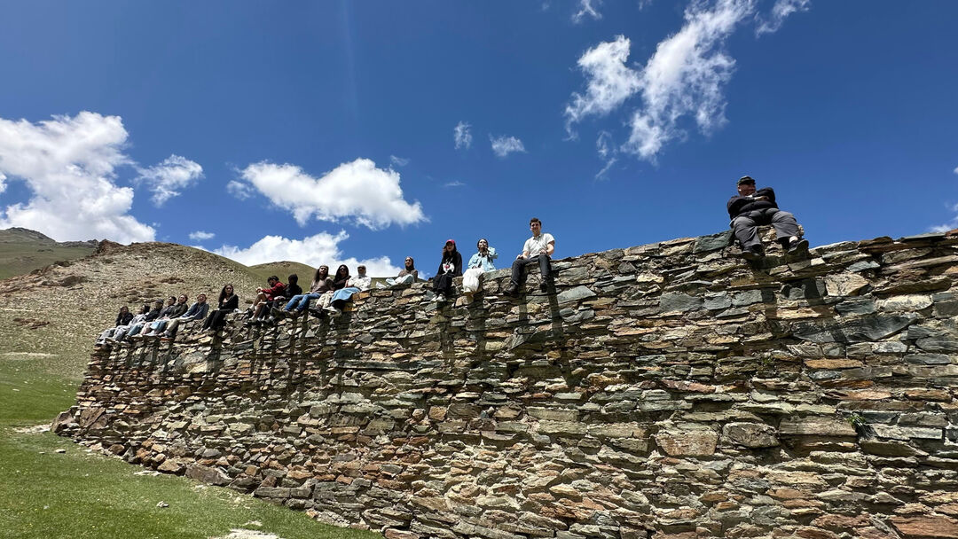 A large group of students, smiling at the camera while sitting on a large rock looking down at the camera from height with greenery and mountains in the background