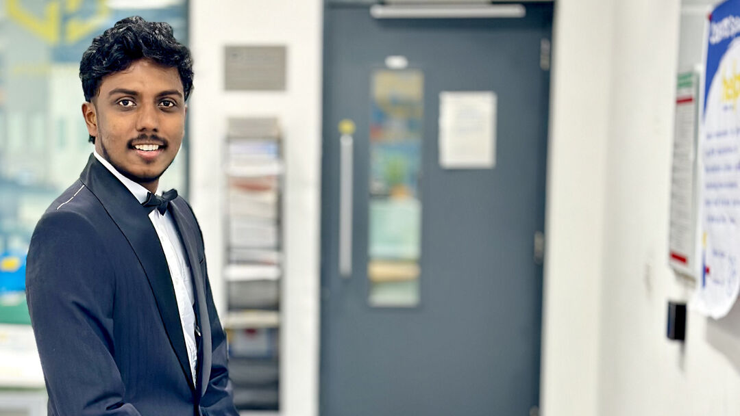 : Student dressed smartly in a blue suit and bow tie, smiing at the camera inside the University building.