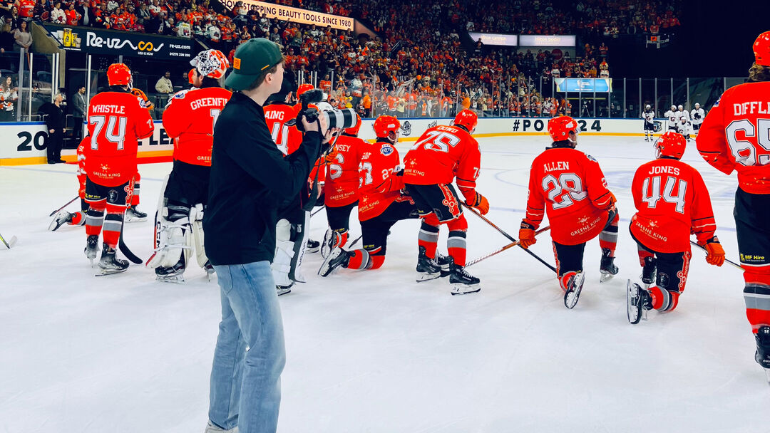 Student wearing a black top, backwards cap and blue jeans with their back to the camera holding a camera, taking a picture of ice hockey players wearing red tops with their backs to the camera on the ice