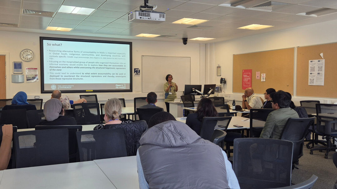 A person wearing a green jumper stands in front of a classroom with people while presenting 