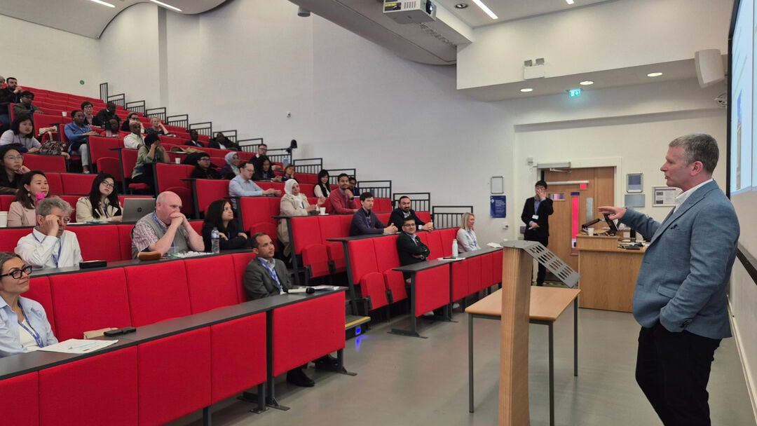 A person wearing a blue jacket and black jeans, talking and facing a audience in a lecture theatre, with red seats. 
