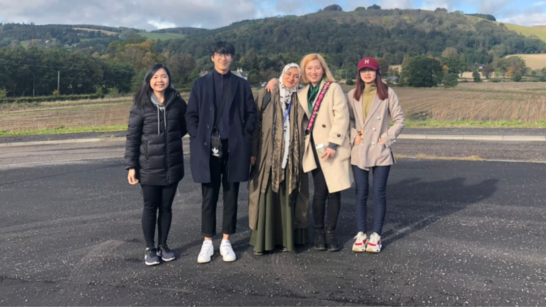 5 students, standing and smiling for the photograph, they are outside with a hills and farms in the background.