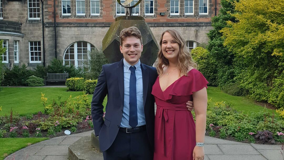 Two people in formal clothing stand in a neat quad in front of a large stone building.
